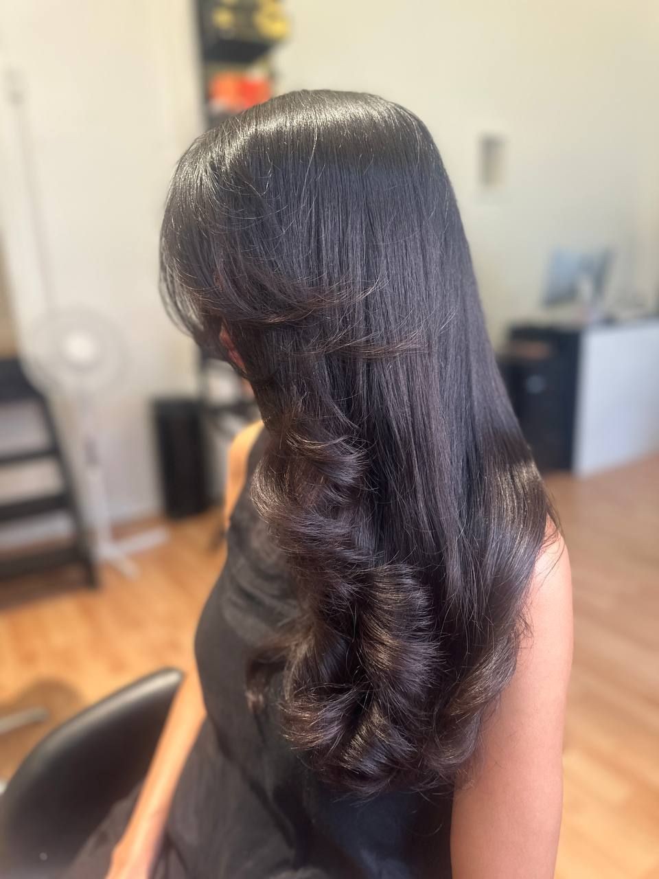 Woman with long, shiny, black hair styled in loose curls sitting in a salon chair.